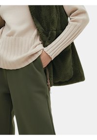 Olijfgroene fleece-vest over een beige geribbelde trui, met donker groene broek. Opvallende details zijn een ritssluitingzak en geribbelde boorden.