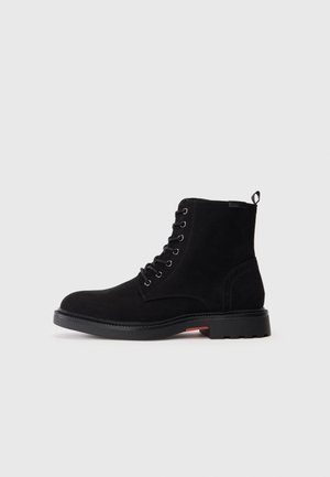 Botas de tornozelo em camurça preta, com bico redondo, atacadores à frente, e sola de borracha com acabamento texturizado e um detalhe vermelho no calcanhar.