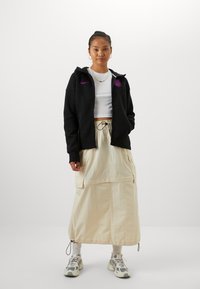 Melnš kapuce krekls ar purpura logo, balts īss tops un bēšs kravas svārki ar priekšējām kabatām un cilpu. Nēsāts ar rakstainām kedām.