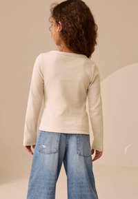 Helles beige, geripptes Langarmoberteil kombiniert mit weiteren blauen Jeans. Das Oberteil hat einen leicht ausgeschnittenen Rundhalsausschnitt.