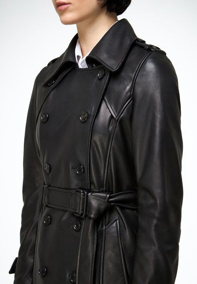 Femme portant un trench-coat en cuir noir à double boutonnage avec ceinture et col large par-dessus une chemise blanche.