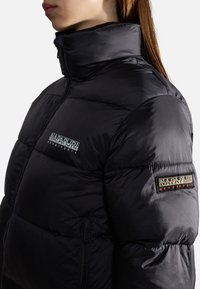Personne portant une veste matelassée noire à col montant avec les logos Napapijri Geographic sur la poitrine et le haut de la manche sur fond blanc.