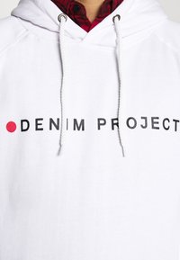 Balta gobtuvė su raišteliu, ant kurios yra juodos ir raudonos spalvos užrašas "DENIM PROJECT." Medžiaga atrodo lygi, su kasdienišku kirpimu.
