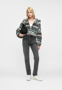 Mikina s kapucňou na zips z flísu s geometrickým vzorom v odtieňoch šedej, ružovej a krémovej, skombinovaná s vysokým pásom šedými džínsami a bielymi teniskami. Drží čiernu tašku.