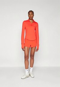 Heledat oranž värvi pikkade varrukatega lukuga särk pöidlaavadega, sobitatud lühikestega. Kandes valgete spordijalatsite ja keskmise pikkusega sokkidega.