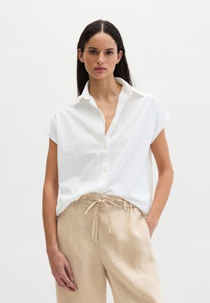 Frau mit langen dunklen Haaren, die ein weißes Kurzarm-Hemd mit Knöpfen und beige Hosen mit Kordelzug vor einem schlichten Hintergrund trägt.