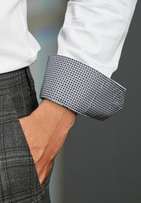 Camicia bianca testurizzata con un polsino decorato in blu e bianco. Il polsino presenta un motivo circolare. Pantaloni a quadri grigi visibili. Mano in tasca.