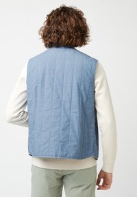Blauer gesteppter Weste mit einer strukturierten Oberfläche, ärmellosem Design und seitlichen Taschen. Über einem hellen Langarmshirt getragen.