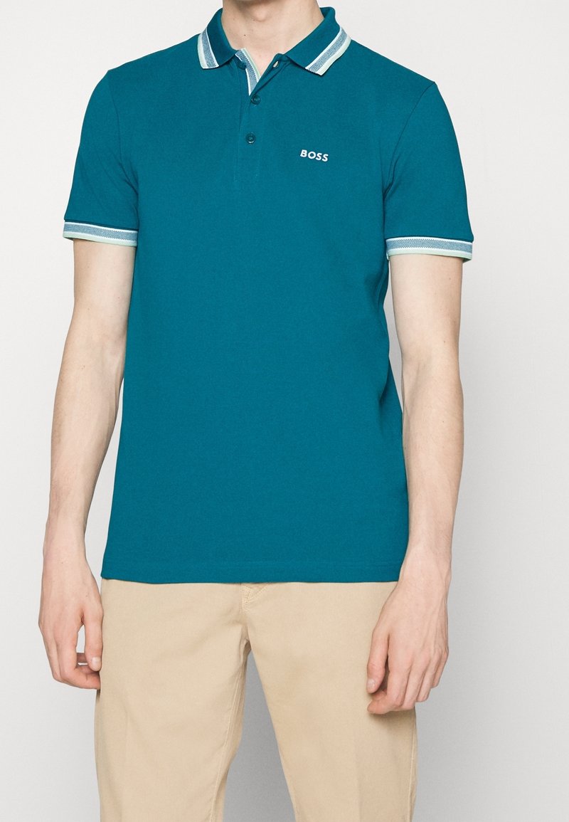 Homme portant un polo bleu sarcelle avec un col et des bords de manches rayés blanc et bleu clair, pantalon beige, chemise avec une étiquette BOSS sur la poitrine.