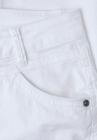 Gros plan d’un jean en denim blanc montrant la poche avant, le passant de ceinture et le détail du rivet métallique sur le tissu.
