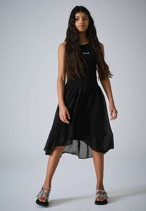 Juoda be rankovių suknelė su prigludusiu liemeniu ir laisva, iki kelių siekiančia sijono dalimi. Dėvima su pilkais sandalais, kuriose yra sidabro spalvos metalinės detalės.