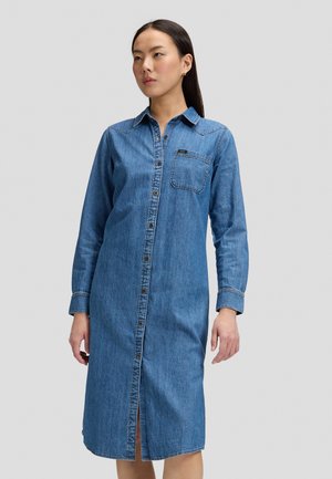 Femme portant une robe chemise en denim bleu à manches longues, longueur genou, avec une poche poitrine, debout sur un fond gris clair uni.