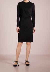 Svart långärmad klänning med tvinnad frontdetalj, mjukt jersey tyg, knälång fåll, tillsammans med svarta klackskor med ankelsnören.