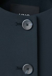 Gros plan d'un manteau en tissu noir avec deux grands boutons noirs cousus, attachés à travers des boutonnières horizontales.
