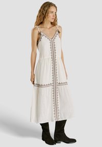 Žena s vlnitými vlasmi v bezrukávových bielych midi šatách s geometrickou výšivkou a čiernymi čižmami po kolená, stojaca a pozerajúca sa doprava.