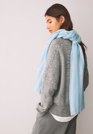 Femme aux longs cheveux foncés porte un pull gris et une écharpe rayée bleu clair drapée sur les épaules, regardant vers le bas sur un fond neutre.