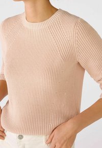 Kobieta w jasnoróżowym swetrze z fakturowanej dzianiny z rękawami do łokci włożonym w białe spodnie, ręka oparta na biodrze.
