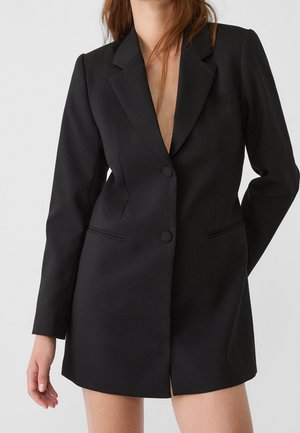 Nainen, joka pukeutunut pitkään mustaan bleiserimekkoon, jossa on lovinmuotoiset kaulukset, kaksi etunappia ja läppätaskut, yksinkertaista vaaleaa taustaa vasten.