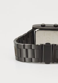 Montre en acier inoxydable noir avec un boîtier rectangulaire, une lunette polie, une finition mate sur le bracelet et deux boutons sur le dessus.
