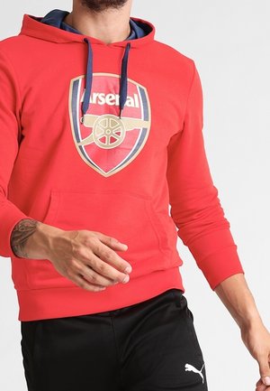 Mand iført rød Arsenal-hættetrøje med marineblå snore og sorte Puma-bukser, stående mod en ensartet lys baggrund.