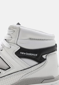 White high-top sneaker with black accents, featuring leather and mesh materials, a padded collar, and perforated detailing on the side.