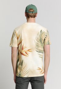 Korte mouwen, beige T-shirt met een tropische palmbladenprint in groene en oranje tinten, gedragen met een groene pet. Zachte stof met een relaxte pasvorm.