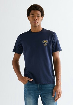 Marineblaues Baumwoll-T-Shirt mit Rundhalsausschnitt, kurzen Ärmeln und einem kleinen gelb-weißen Logo auf der linken Brust. Kombiniert mit blauen Jeans aus Denim.
