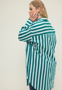 Camisa de rayas verticales verdes y blancas con un ajuste relajado, mangas largas y dobladillo curvado, hecha de una tela suave.