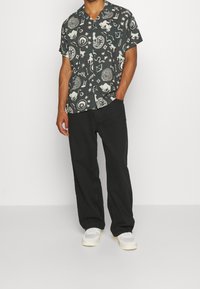 Camicetta nera a maniche corte con motivi bianchi astratti, abbinata a pantaloni neri e sneakers bianche. Tessuto in cotone, vestibilità rilassata.
