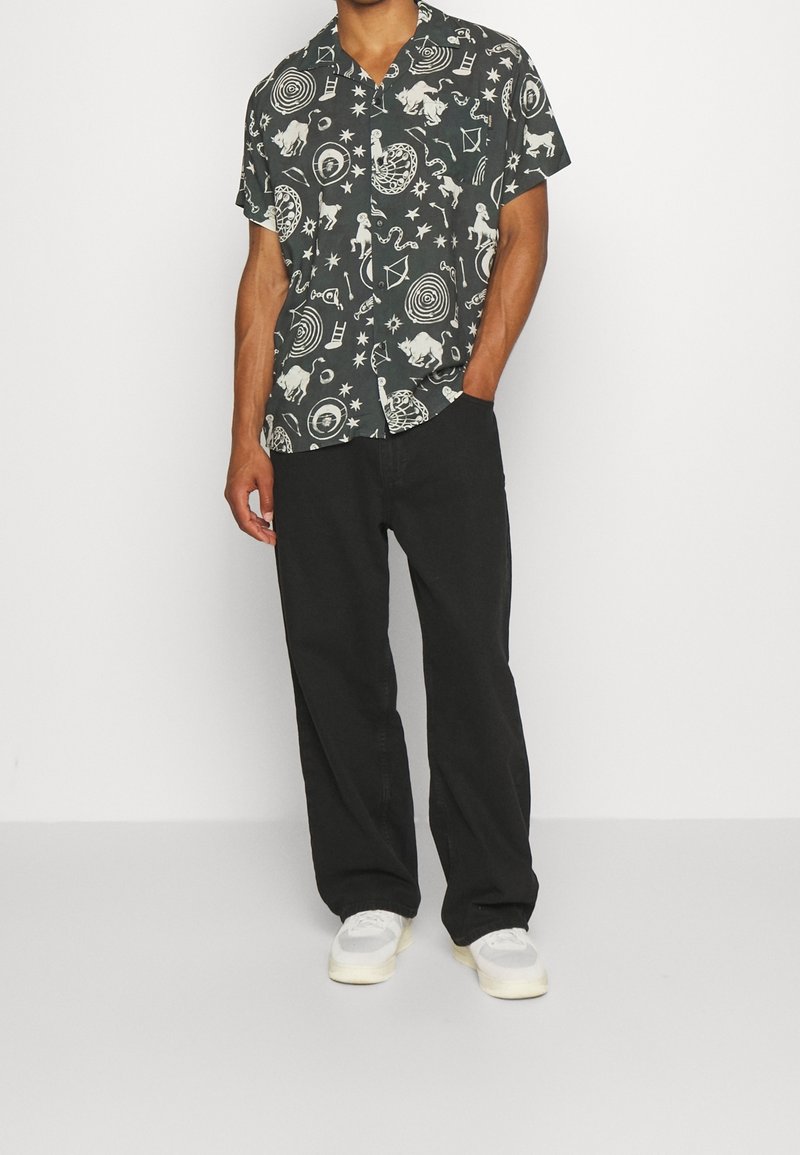 Camicetta nera a maniche corte con motivi bianchi astratti, abbinata a pantaloni neri e sneakers bianche. Tessuto in cotone, vestibilità rilassata.