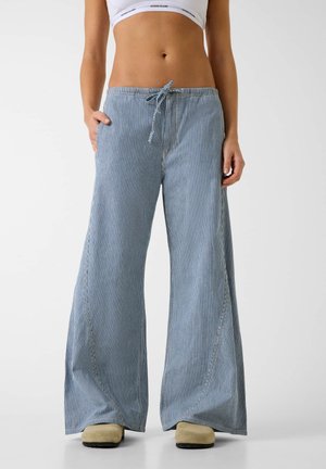 Kobieta ubrana w jasnoniebiesko-białe, pionowo paskowane spodnie o szerokich nogawkach z wiązanym paskiem w talii oraz beżowe buty, stojąca na jednolitym tle.