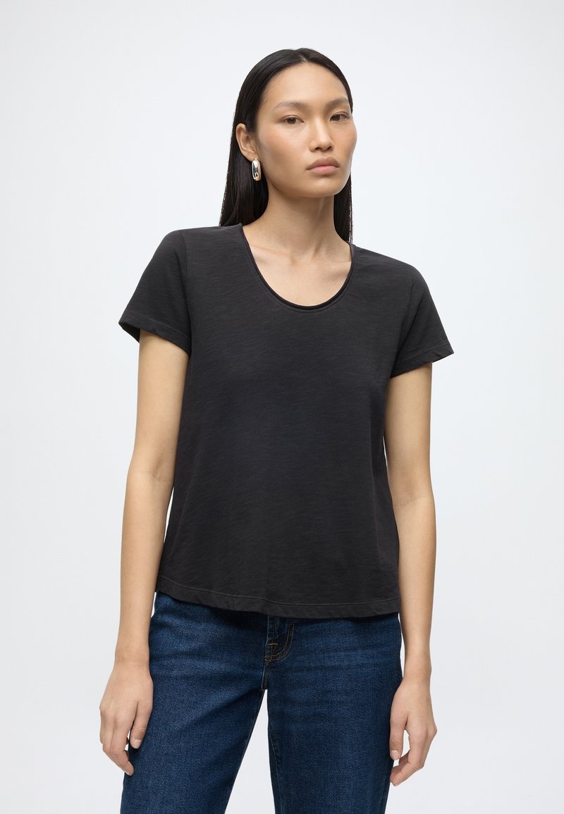 Jeune femme aux longs cheveux foncés portant des boucles d'oreilles en argent en forme d'anneau, un t-shirt noir uni à manches courtes et un jean bleu, debout devant un fond blanc.