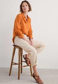 Oranžová lněná košile na knoflíky s dvěma kapsami na hrudi, spárovaná se světle béžovými kalhotami. Hnědé sandály na pásky. Sedící na dřevěném stoličce.