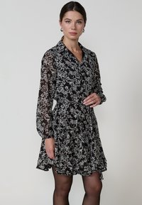 Rochie neagră cu flori, cu mâneci lungi din material transparent, guler, talie strânsă și fustă în straturi. Prezintă modele florale albe pe un fundal negru.