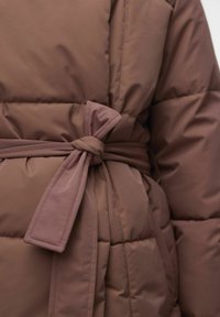 Brun pufferjakke med belte, med kviltede seksjoner, en glatt tekstur og en sløyfedetalj i midjen for ekstra definisjon.