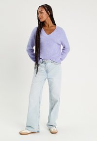 Lichtpaarse gebreide trui met een V-hals, gecombineerd met lichtblauwe wijde jeans en beige sneakers. Lange gevlochten haren. Eenvoudige, casual outfit.