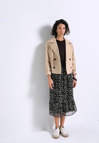 Femme portant une veste beige, un haut noir, une jupe midi à motifs noir et blanc, et des baskets blanches, debout contre un mur blanc.