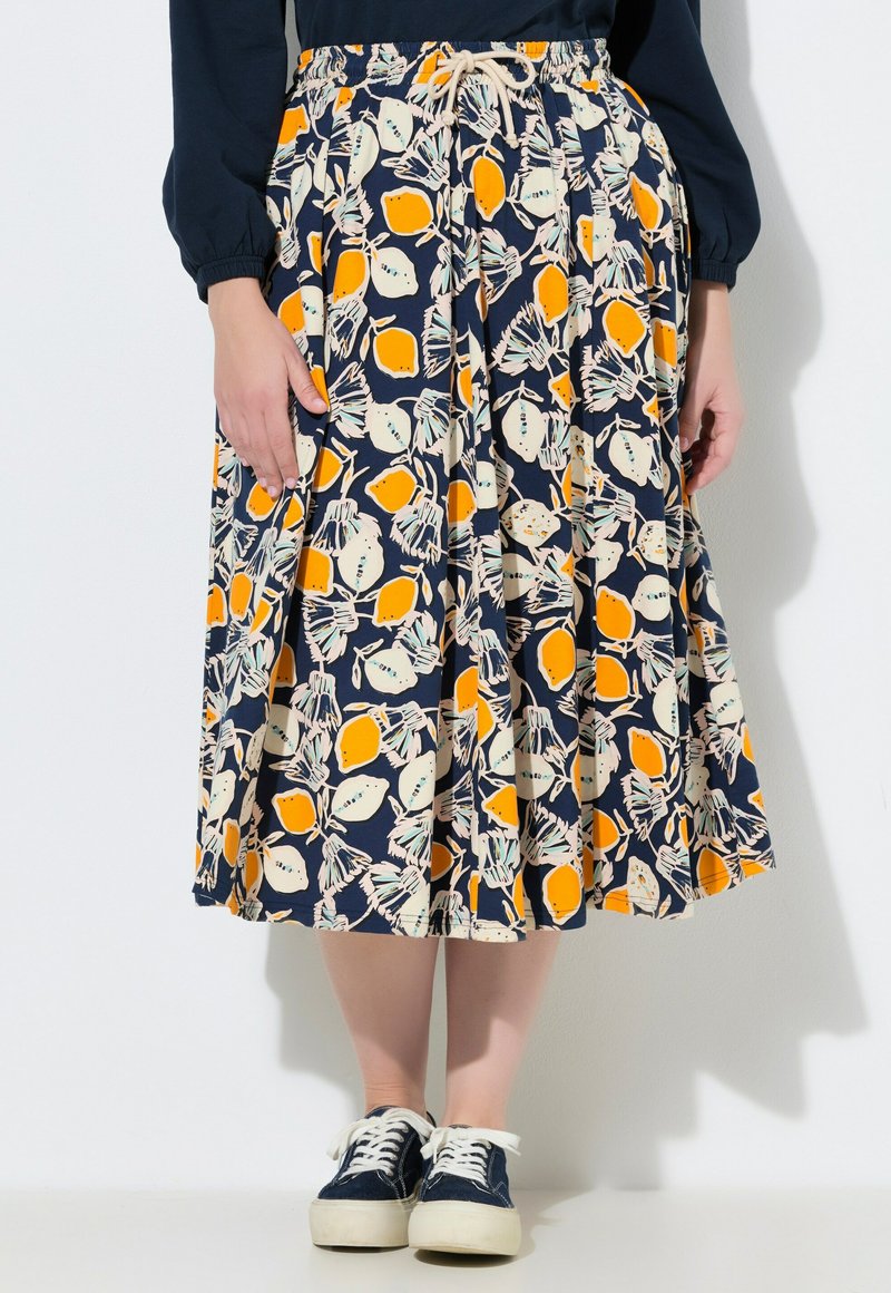 Jupe midi bleu marine avec imprimé citrons orange et crème, taille froncée avec cordon de serrage, portée avec des baskets bleu marine et un haut à manches longues bleu marine.