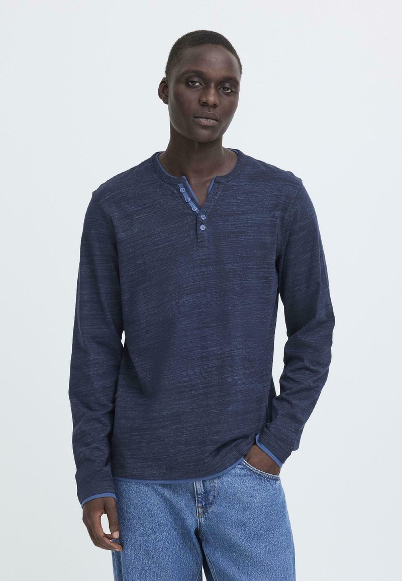 Marineblå langermet henley-skjorte med strukturert stoff, tre-knappers placket, og kontrastfarget blå krage og mansjetter. Brukt med lyse denimjeans.