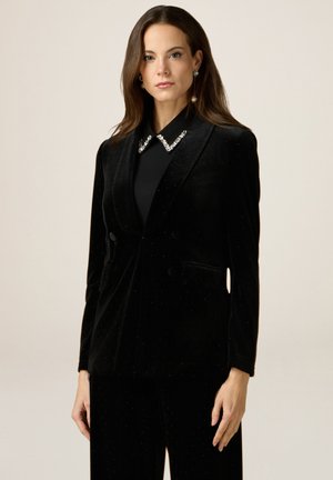 Blazer de terciopelo negro con un diseño de doble botonadura, cuenta con un cuello adornado con tachuelas y bolsillos. Se combina con pantalones a juego en una textura similar.