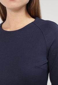 Dunkelblaues Oberteil mit rundem Ausschnitt und dezenten Schulternähten, aus glattem, elastischem Stoff gefertigt. Es hat eine figurbetonte Silhouette.