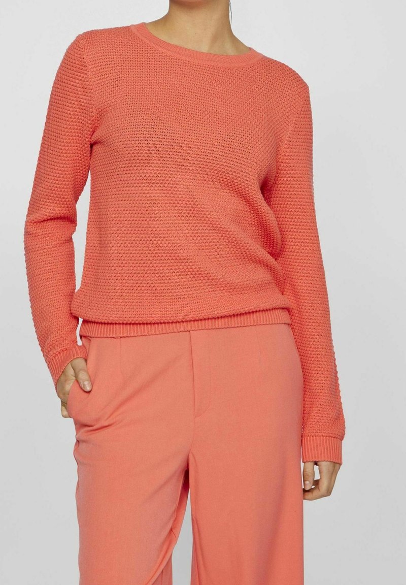 Femme portant un pull tricoté texturé corail et un pantalon corail assorti, main dans la poche du pantalon, sur un fond uni gris clair.
