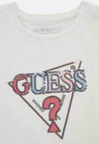 Camiseta blanca de algodón orgánico con un logo en forma de triángulo decorado con lentejuelas que dice "GUESS" en azul y rosa, y un acento de signo de interrogación en rosa.