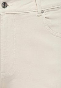 Close-up van off-white denim jeans waarop de voorkant zak, riemlus en zilveren knoop met gestikte details te zien zijn.