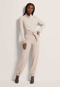Hellbeige gerippter Rollkragenpullover mit langen Ärmeln; hellbeige Hose mit tailliertem Bund und schmal zulaufenden Beinen; kombiniert mit dunkelbraunen Stiefeletten.