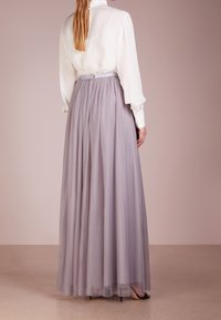 Femme portant un chemisier blanc à manches longues et une jupe plissée légère violet clair jusqu'au sol avec des talons, se tenant devant un fond uni.