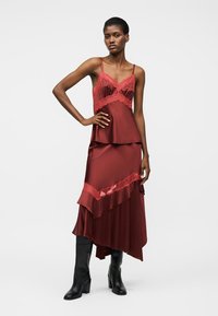 Žena v bezrukávových bordových saténových šatách s čipkovými detailmi, v kombinácii s čiernymi čižmami nad kolená na vysokom opätku, stojaca s rukou na páse.