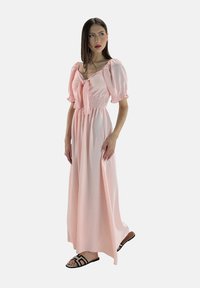 Langes rosa Kleid mit Puffärmeln, geraffter Taille und Schleife an der Vorderseite. Leichter Stoff, glatte Textur, lockere Passform. Kombiniert mit schwarzen Sandalen.