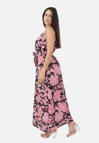 Donna con lungo capelli castani, che indossa un abito maxi senza maniche floreale nero e rosa e sandali trasparenti con tacco a blocco, in piedi di lato.