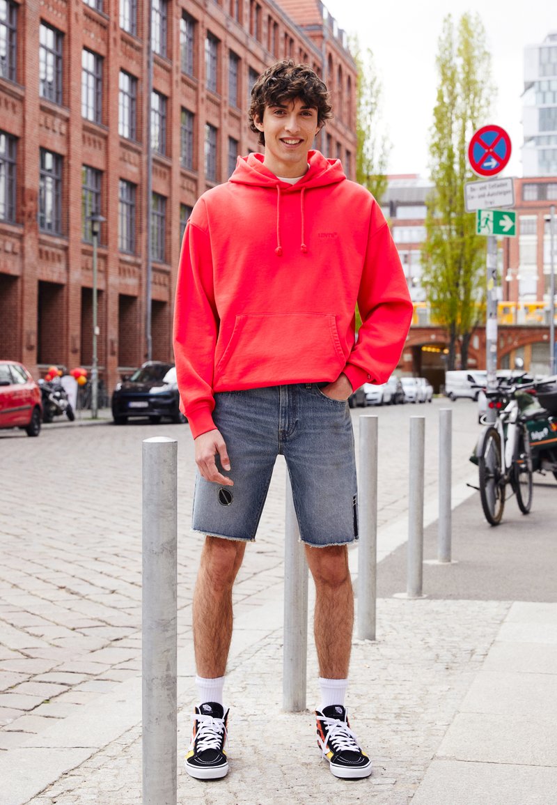 Neon roze hoodie met een kangoeroezak, gecombineerd met distressed denim shorts en zwarte sneakers, voor een achtergrond van een stadsstraat.